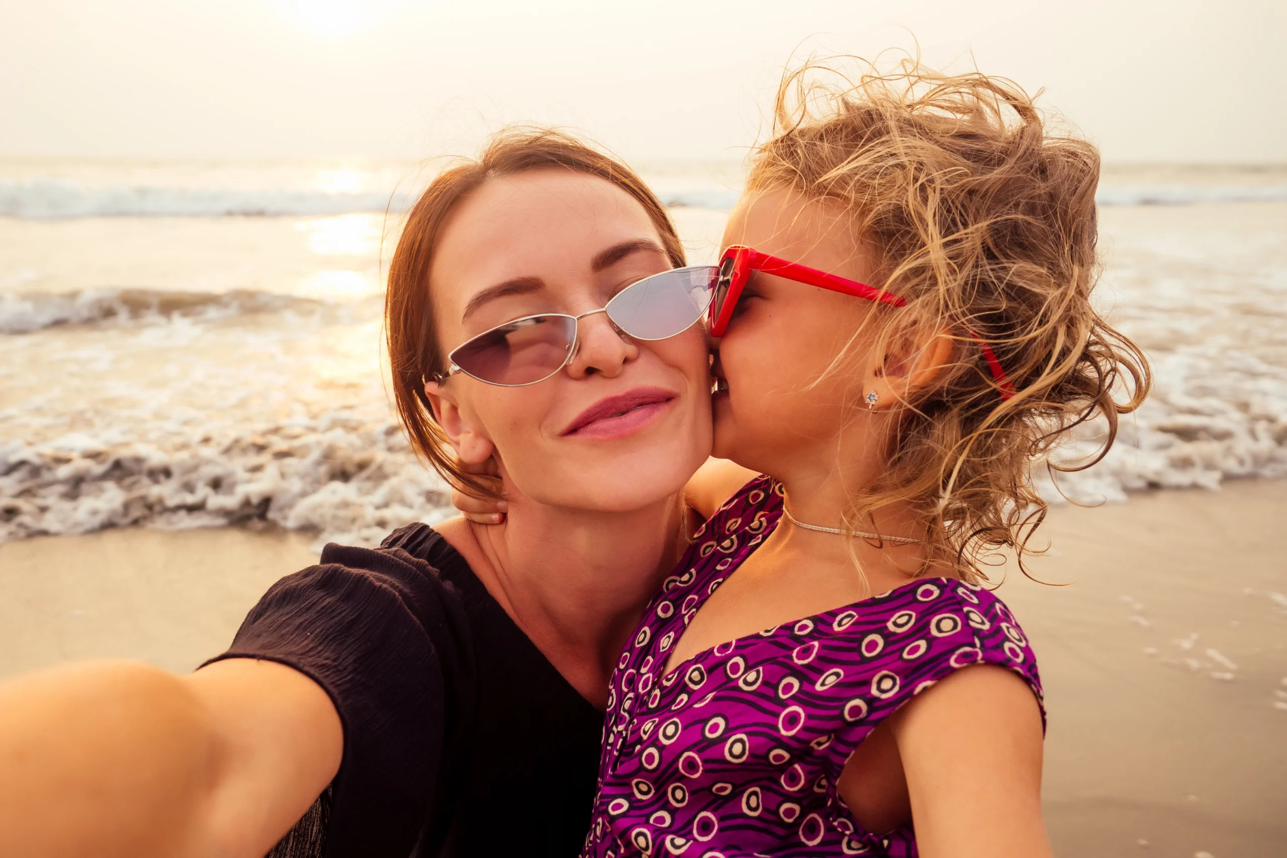 A selfie of a smiling woman and child, both wearing sunglasses at the beach. The child is kissing the woman on the cheek.